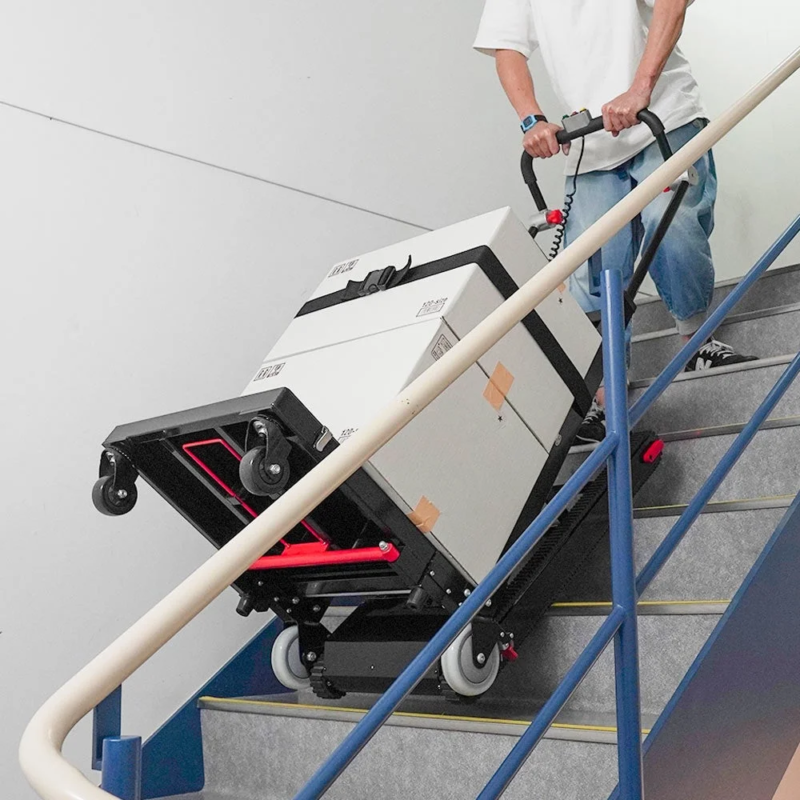 階段を上る人物が、電動式の階段昇降台車を使って大きな白い荷物を運んでいる様子