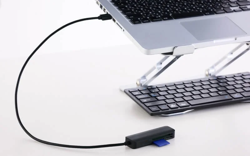 ノートパソコンがスタンドに置かれ、USBケーブルで接続されたカードリーダーにSDカードが挿入されています。その下には外付けキーボードがあり、効率的な作業環境が示唆されています。