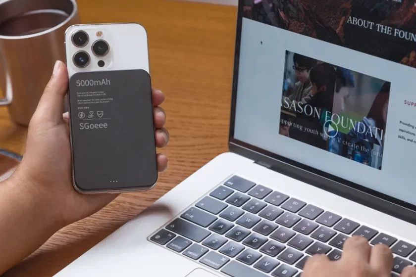 デスクで作業中の人物が、スマートフォンに装着されたモバイルバッテリーを手に持ち、隣のノートパソコンの画面には財団のウェブサイトが表示されています。