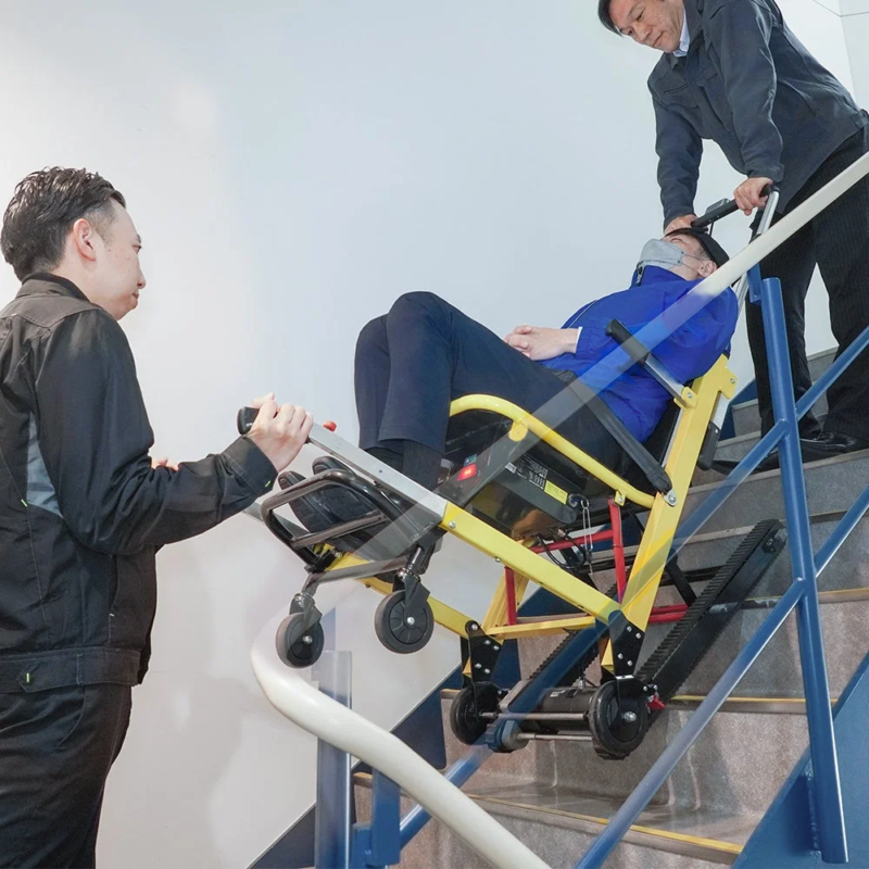 二人の男性が協力して、階段昇降機に乗った人物を階段で安全に移動させている様子