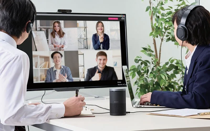 オフィスで働く2人がヘッドホンを着用し、パソコンと大型モニターを使ってオンライン会議に参加している様子です。