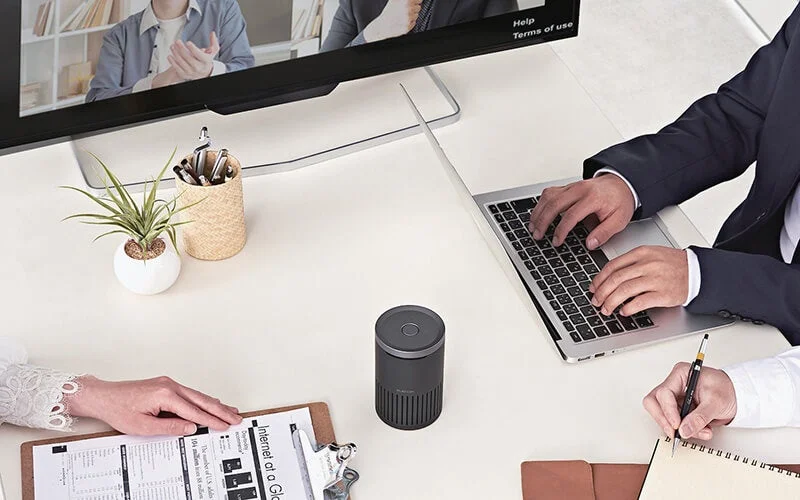 オフィスで複数のビジネスパーソンが会議を行っており、一人はノートパソコンを操作し、別の一人は書類を確認しています。モニターにはオンライン会議の参加者が映し出され、円筒形のデバイスが中央に置かれています。