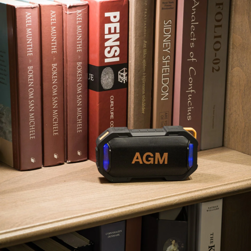 木製の本棚に並べられた様々な本の前に、AGMブランドの黒いポータブルスピーカーが置かれています。