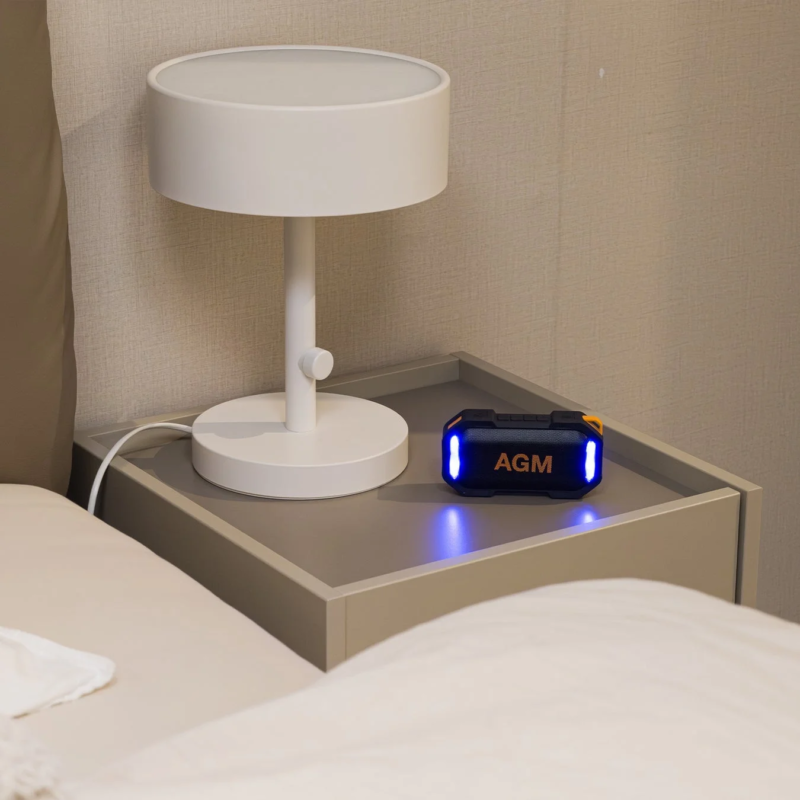 ベッドサイドのナイトテーブルに、シンプルな白いテーブルランプと、青く光る「AGM」のロゴが入った黒いポータブルスピーカーが置かれている様子です。