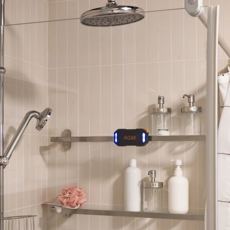 明るいタイル張りの浴室で、シャワーヘッドとハンドシャワーが設置されています。壁の棚には、シャンプーなどのボトルや泡立てネット、そして青く光る「AGM」と書かれたポータブルスピーカーが置かれています。