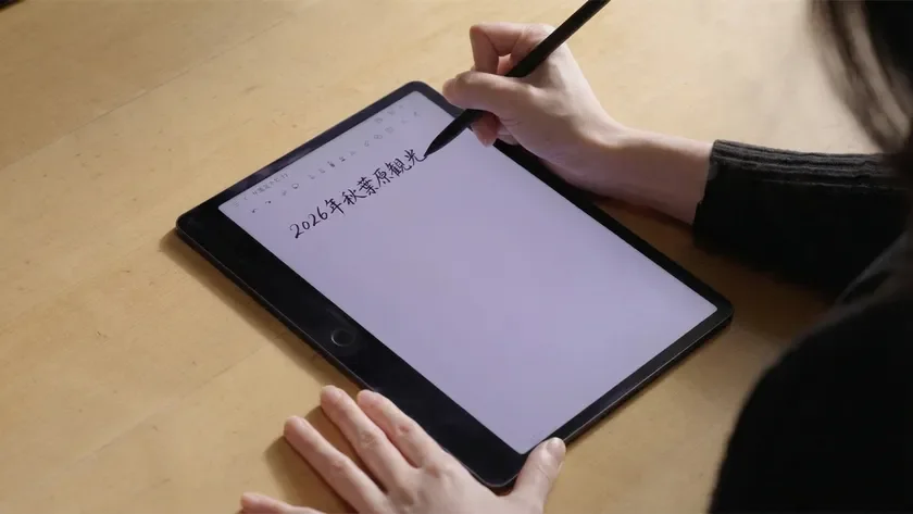 机の上で、一人の人物がデジタルペンを使ってタブレットに「2026年秋葉原観光」と手書きで書き込んでいる様子が写されています。旅行の計画を立てているようです。