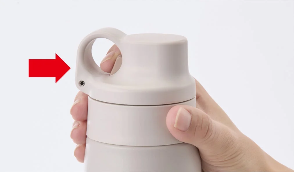 人物がベージュ色の水筒またはタンブラーの蓋にあるハンドル部分を指で持ち、赤い矢印がハンドルの接続部分を指し示している様子を捉えた画像です。