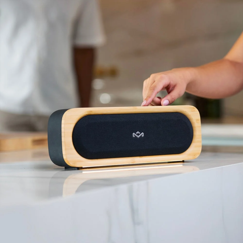 木目調のデザインが特徴的なスピーカーが白いカウンターに置かれ、手が操作しています。背景にはぼやけた人物が写っており、モダンで落ち着いた空間を演出しています。