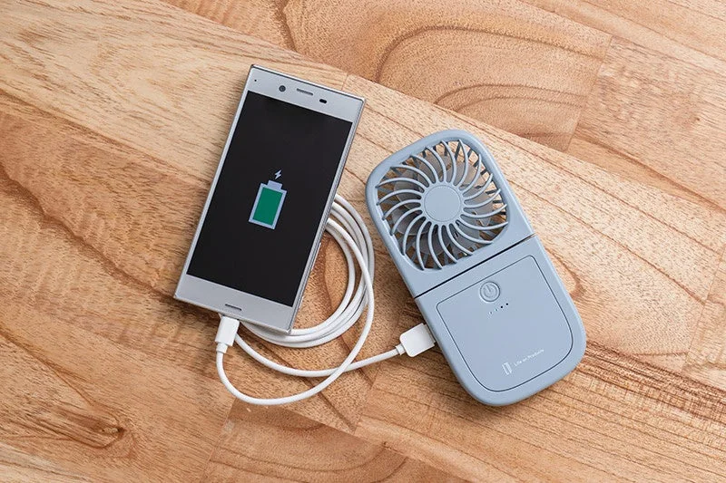 木製の床に置かれたスマートフォンが、ポータブル扇風機兼モバイルバッテリーからUSBケーブルで充電されている様子。