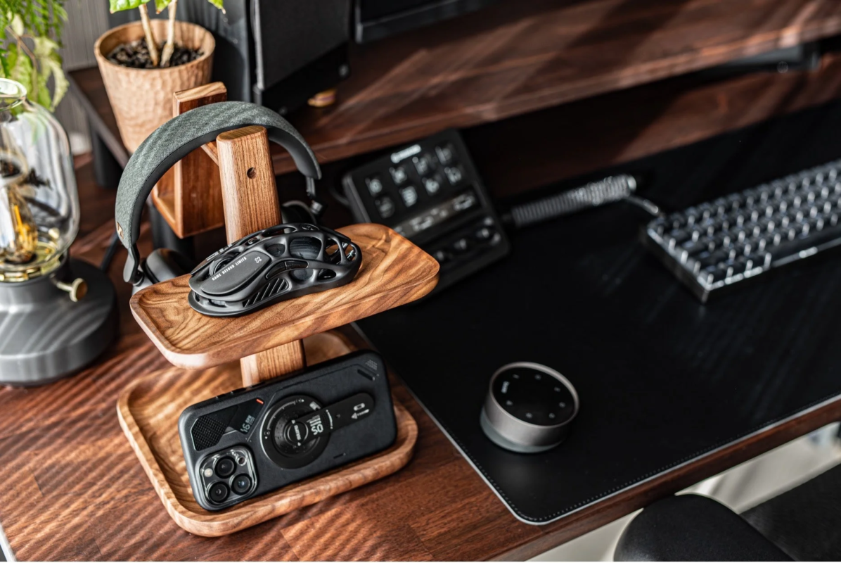 木製のデスクに、ゲーミングマウス、ヘッドホン、スマートフォンが収められた木製スタンドが置かれています。周辺にはキーボードやコントローラー、観葉植物などが配置され、機能的でスタイリッシュなワークスペースを構成しています。