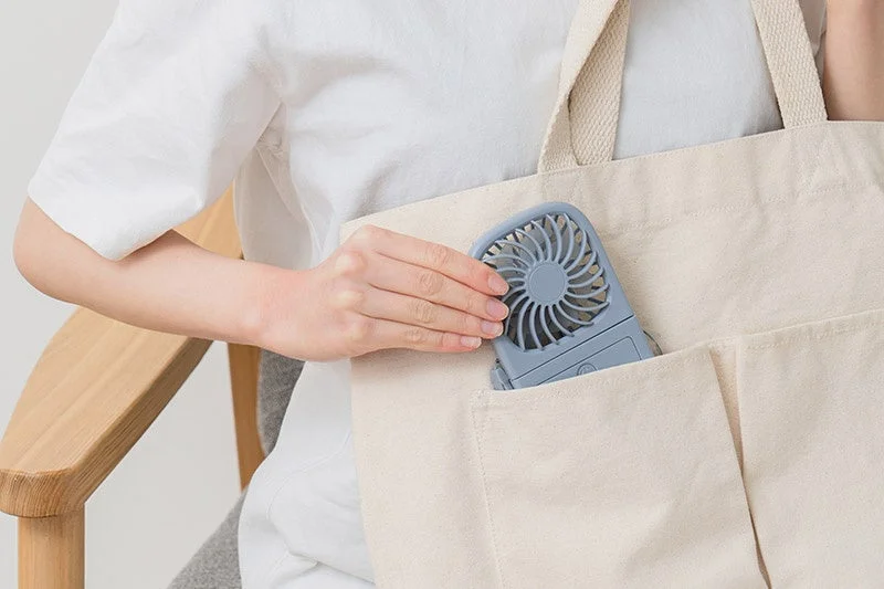 白いTシャツを着た人物が、ベージュ色のトートバッグのポケットに灰色のポータブル扇風機を収納している様子です。