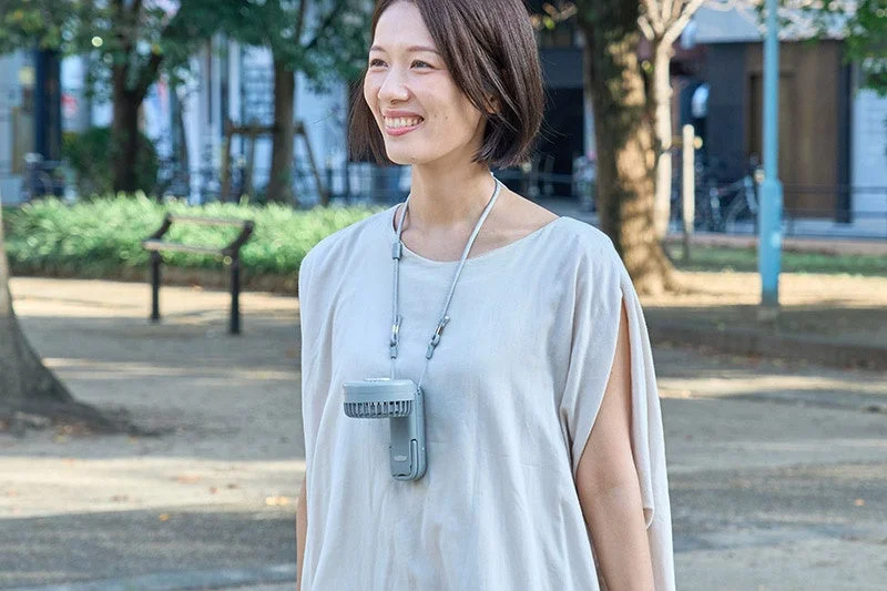 屋外で、笑顔の女性が首から携帯型のネックファンを下げている様子