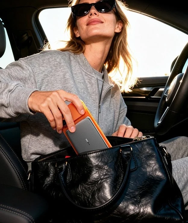 サングラスをかけた女性が車内で、オレンジ色のケースに入ったスマートフォンを黒いハンドバッグにしまっている様子