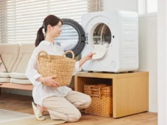 一人の女性が、リビングのような明るい空間で、衣類乾燥機に洗濯物を入れている様子です。手元には編み込みのかごがあります。