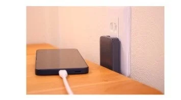 スマートフォンが木製の台の上に置かれ、白い充電ケーブルで壁のコンセントに差し込まれた充電器に接続されている様子。