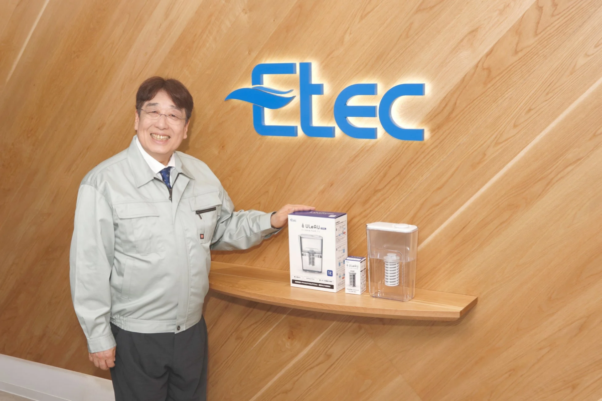 Etecのロゴが入った木目調の壁の前で、笑顔の男性が浄水ポット型浄水器「ULEAU」とそのカートリッジを説明しています