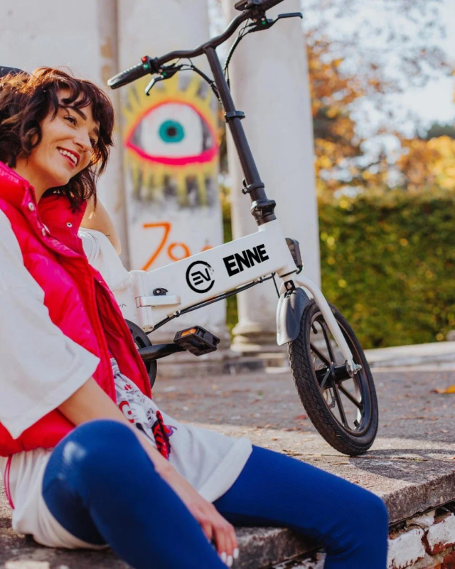 ピンクのベストと青いレギンスを着用した女性が、白い電動折りたたみ自転車の隣で笑顔で座っています。背景にはグラフィティのある壁と秋の木々が見えます。