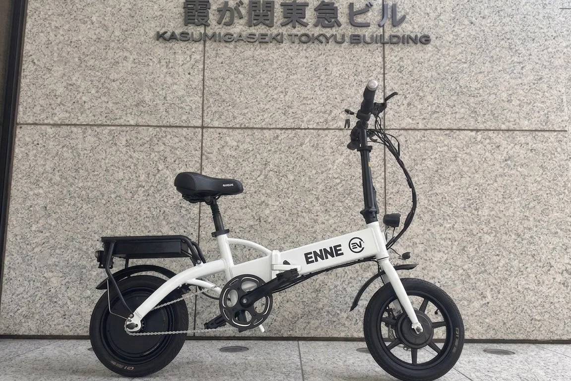 霞が関東急ビルの壁を背景に、白い折りたたみ式電動自転車が置かれています。自転車のフレームには「ENNE EV」の文字が見えます。