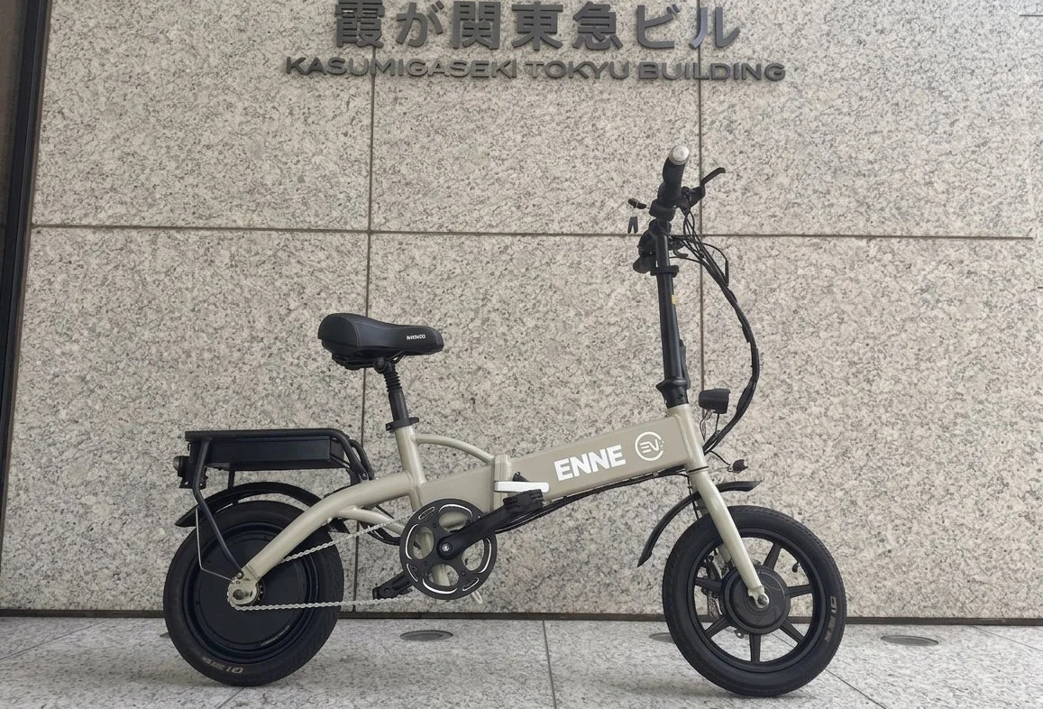 霞が関東急ビルを背景に、ライトグレーの折りたたみ電動自転車が停車している様子を捉えた写真です。都市での移動に便利なe-bikeが主役です。