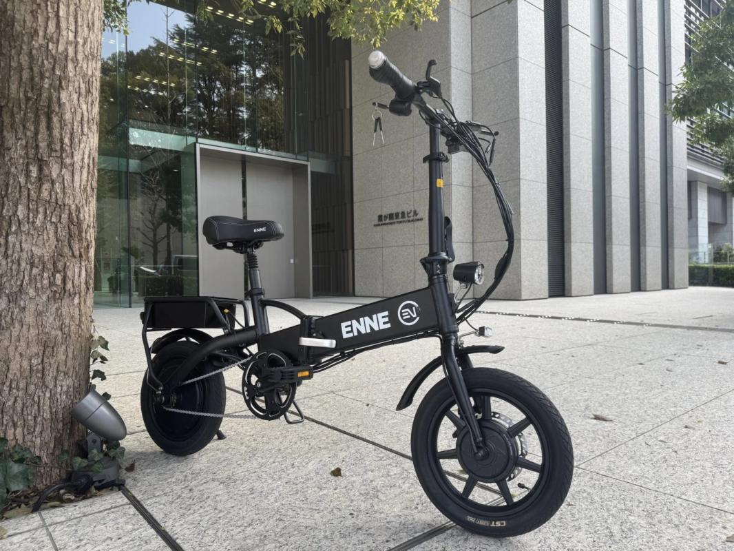 現代的なビルを背景に、黒色の折りたたみ電動自転車が木の下の歩道に停車しています。ENNE EVブランドの車両で、都市の移動手段として注目されます。