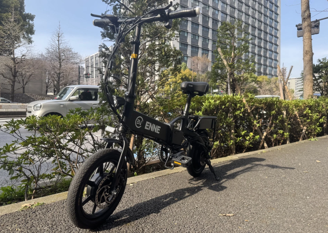 晴れた日に道路脇の植え込みの前に停められた黒い電動アシスト自転車が写っています。奥にはビルや車が見えます。