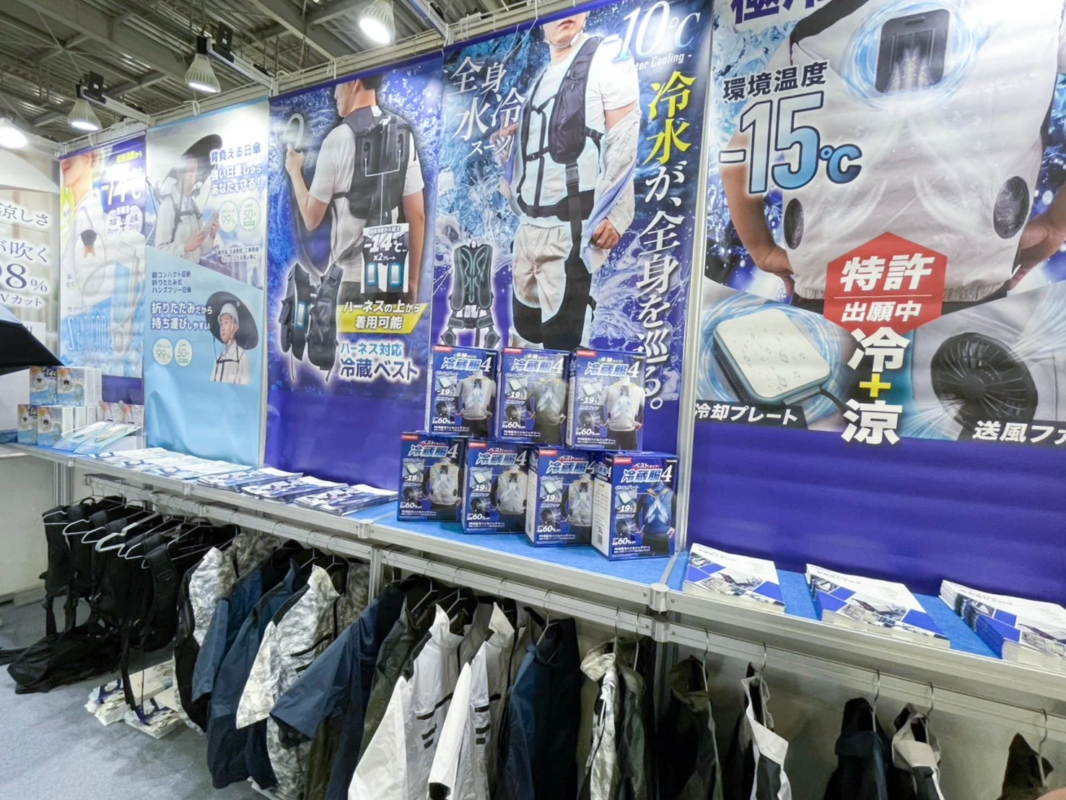 展示会ブースで、水冷ベスト、水冷スーツ、背負える日傘など、暑さ対策や熱中症予防のための様々な冷却グッズが展示されている様子
