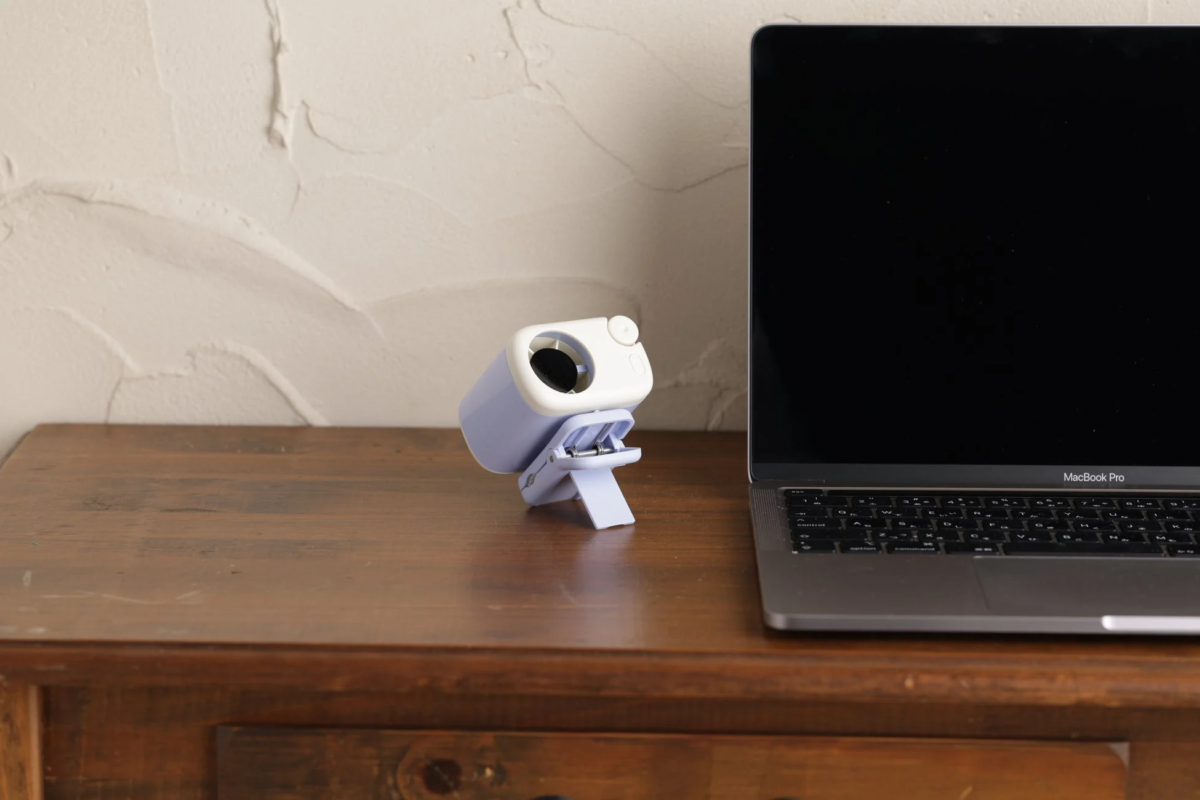 木製のデスクの上に、小型のパープルと白のデバイスが置かれ、その隣にはMacBook Proが配置されています。背景はテクスチャのある白い壁です。