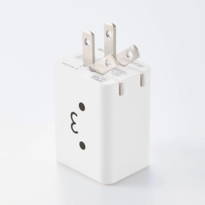 白いシンプルなデザインの電源アダプターで、USタイプのプラグを備えています。側面には可愛らしい顔のイラストが描かれており、機能性と遊び心を兼ね備えています。