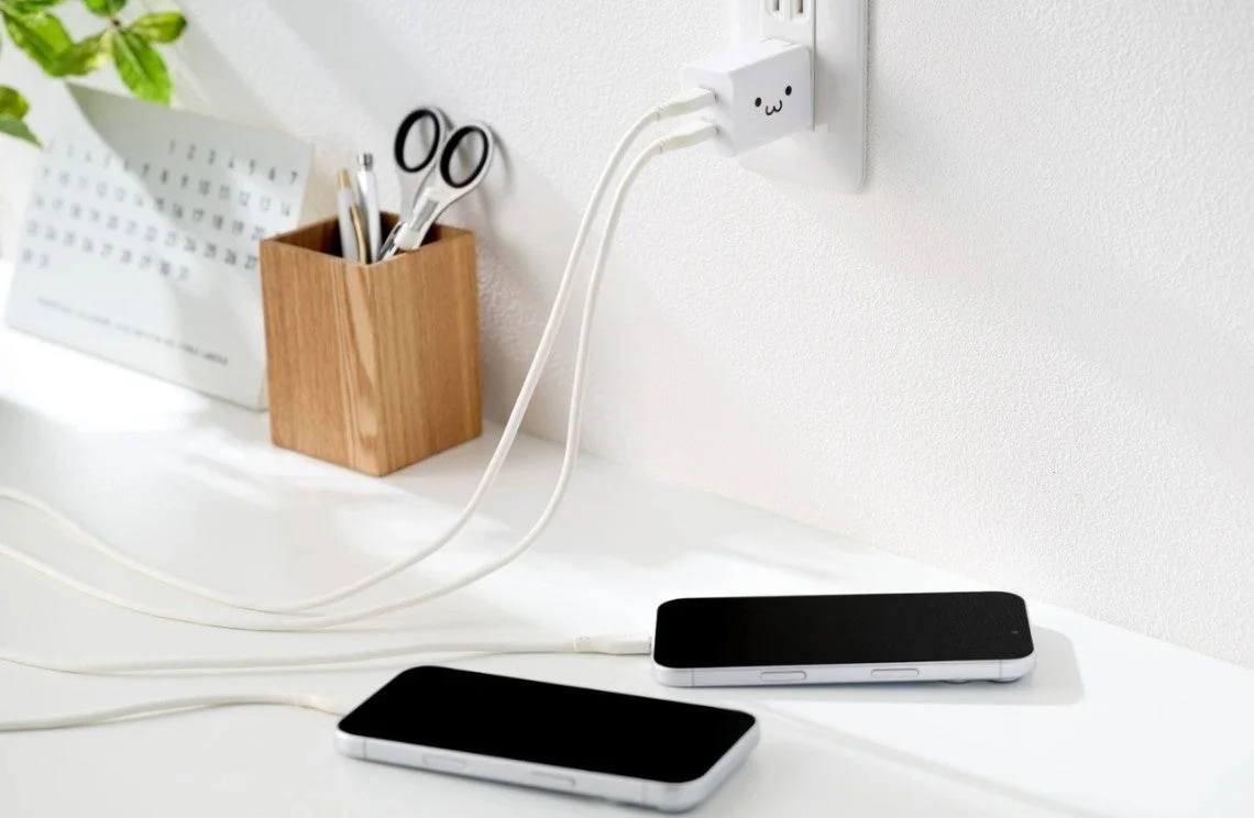 白い壁とデスクを背景に、2台のスマートフォンが充電されている様子を捉えた画像です。コンセントに差し込まれた顔のマークが可愛い電源アダプターから白いケーブルが伸び、清潔感のあるミニマルな空間でデバイスが充電されている日常のシーンが描かれています。